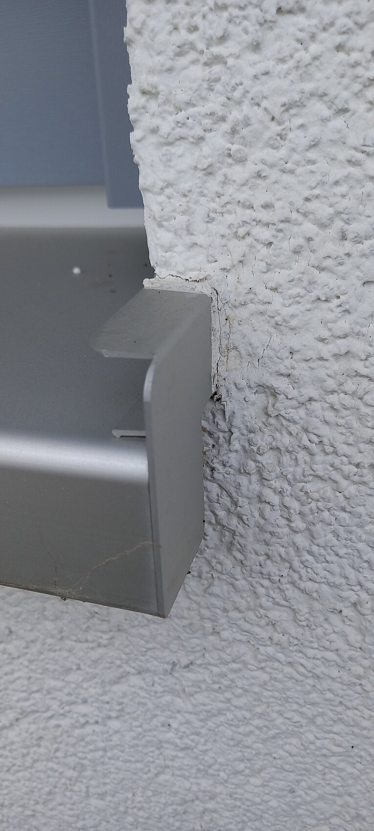 Aussenwand aus rauem weissen Putz mit grauer Metallhalterung und sichtbaren Rissen