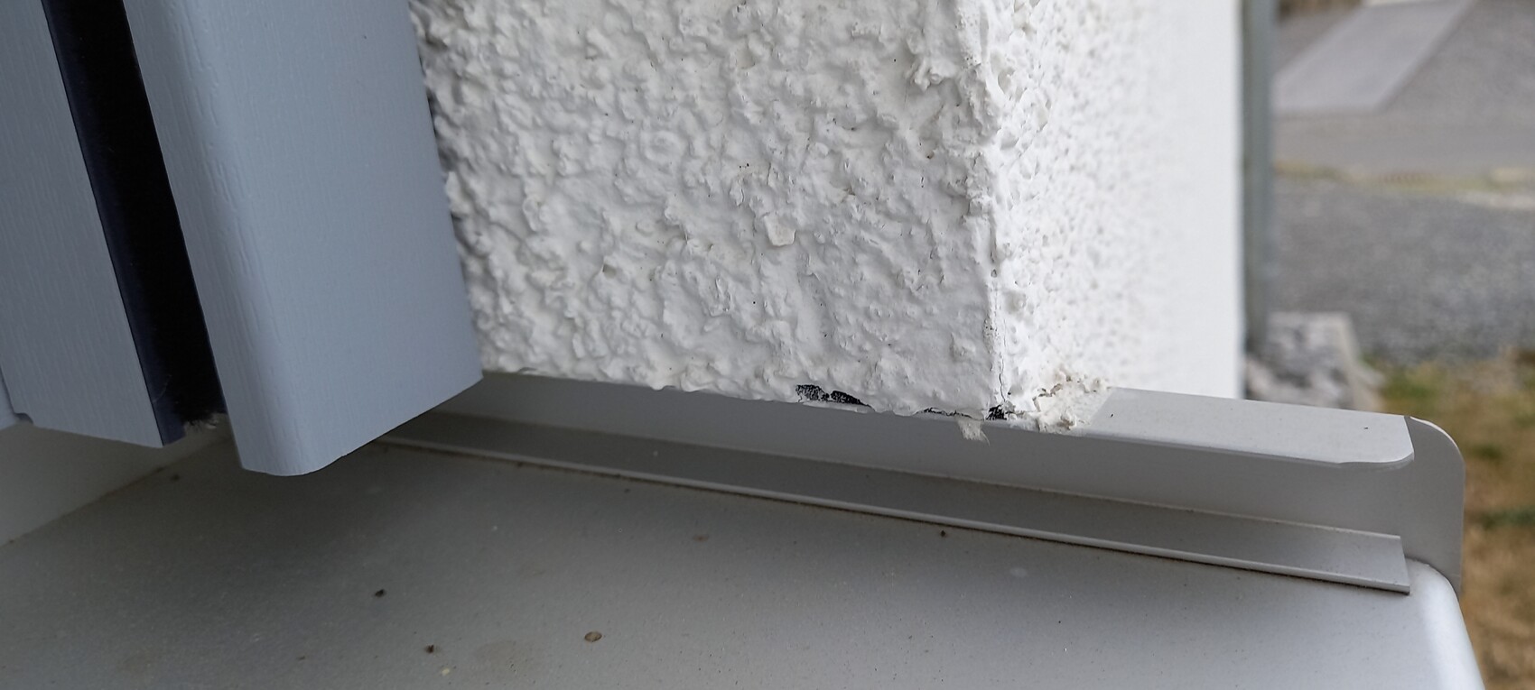 Nahaufnahme einer rauen, weiß verputzten Wand mit Abplatzern unten neben grauem Metall.