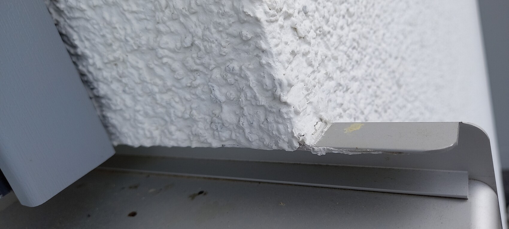 Nahaufnahme einer rauen weißen Putzwand mit grauer Metallauflage und abblätternder Lackierung.