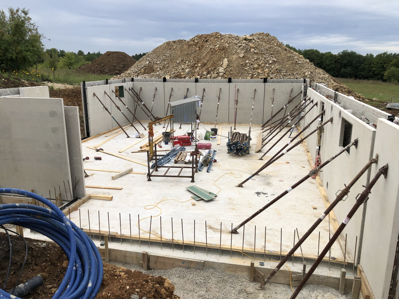 Construction pit with concrete walls, supports, tools, and wooden planks; excavation in the background.