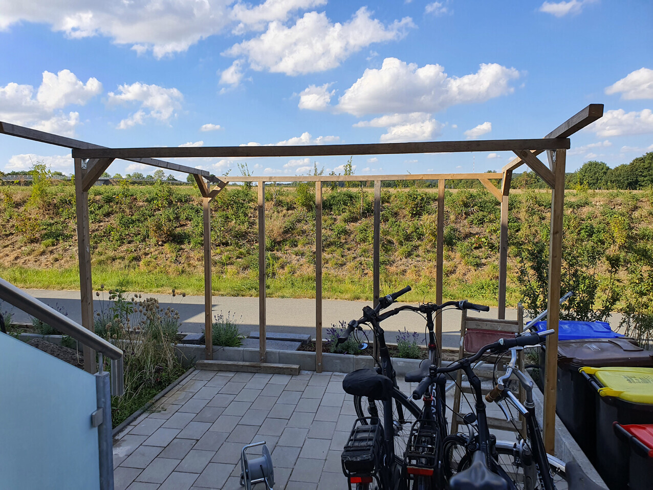 Wooden frame over a paved terrace; several bicycles in front, trash cans to the side.