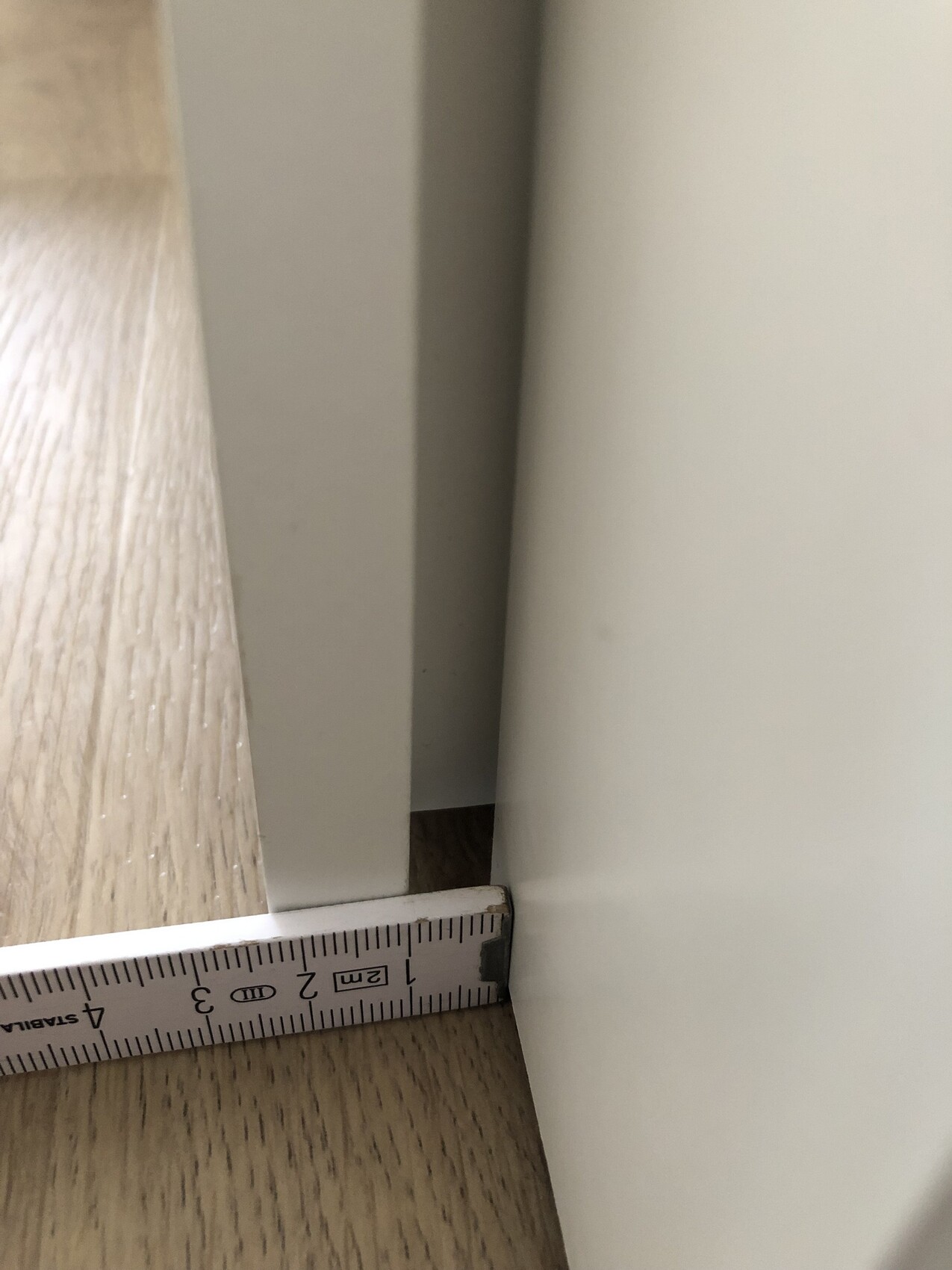Close-up: wooden floor on the left, grey edge, white furniture on the right; tape measure shows approx. 2 cm (1 inch) gap.