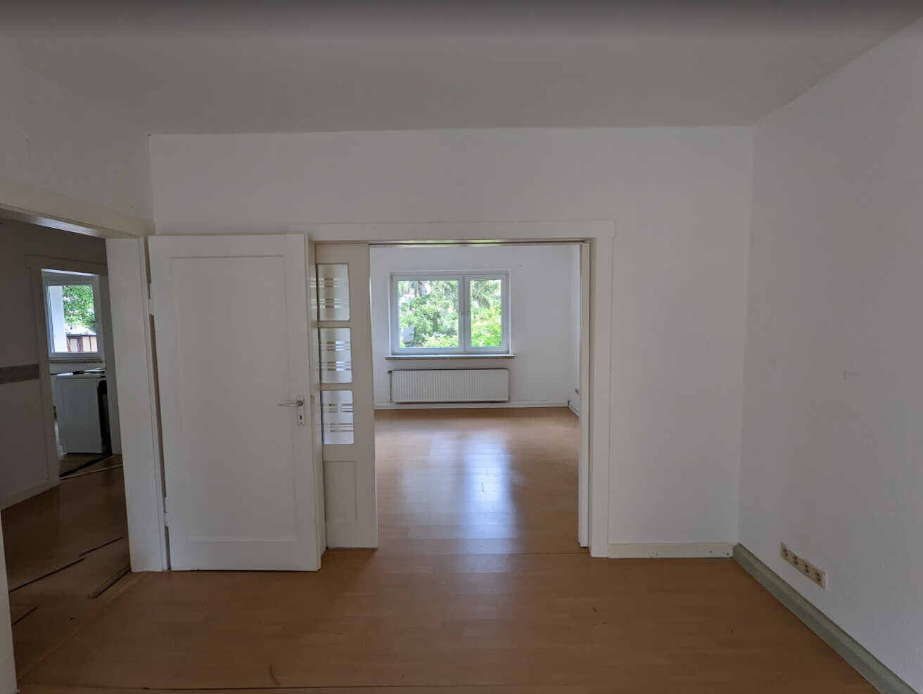 Bright empty living spaces with wooden floor, open door frame and window with garden view.