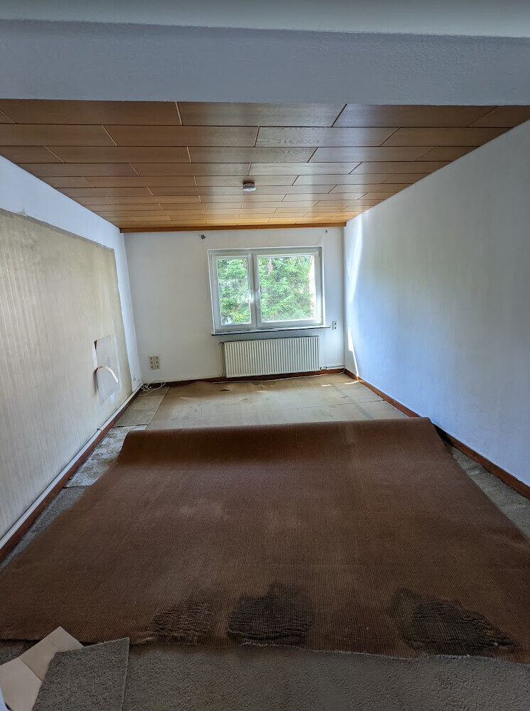 Narrow attic room with wood ceiling, white walls and brown carpet; window with radiator.