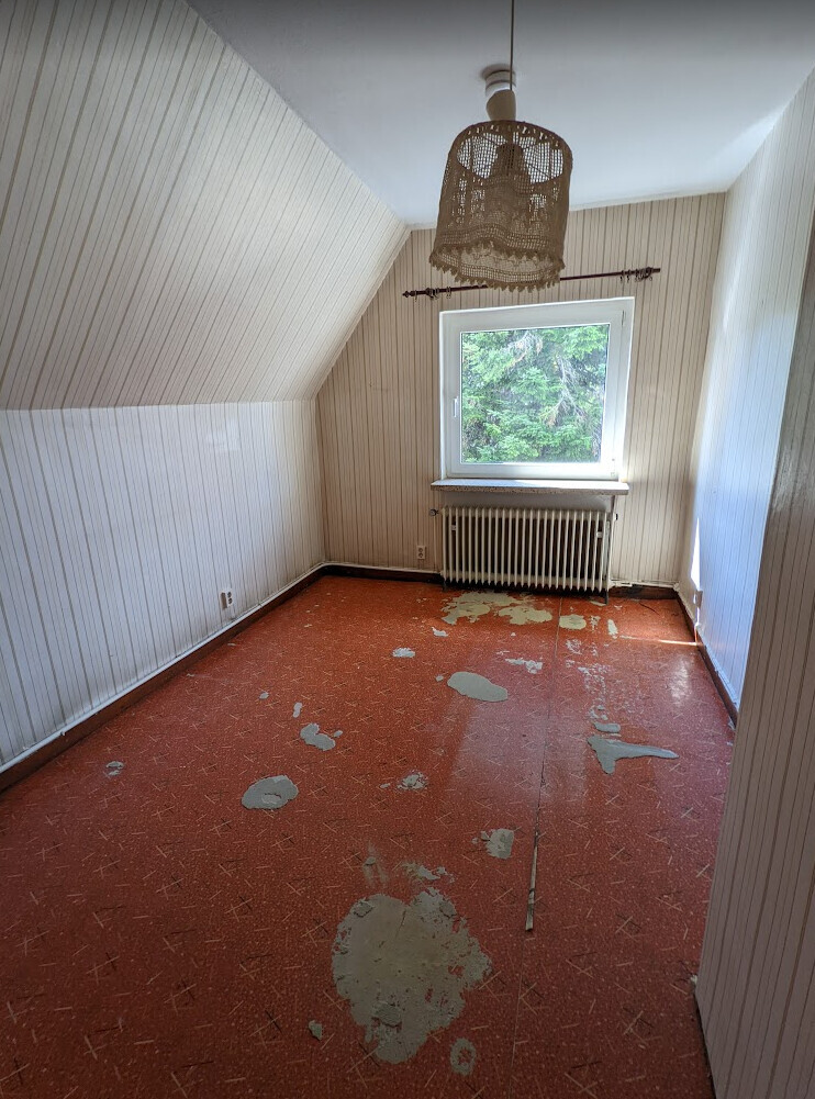 Small, empty room with striped walls, window above radiator, basket lamp, red floor.