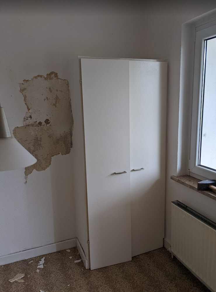 Weißer Zweitüriger Kleiderschrank steht in einem Zimmer mit beschädigter Wand und Fenster.