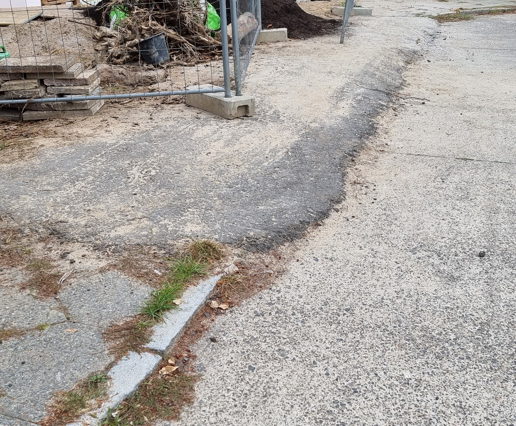 Baustellenbereich: Metallzaun, Holzreste, Paletten; dunkler Asphaltstreifen am Rand der Straße
