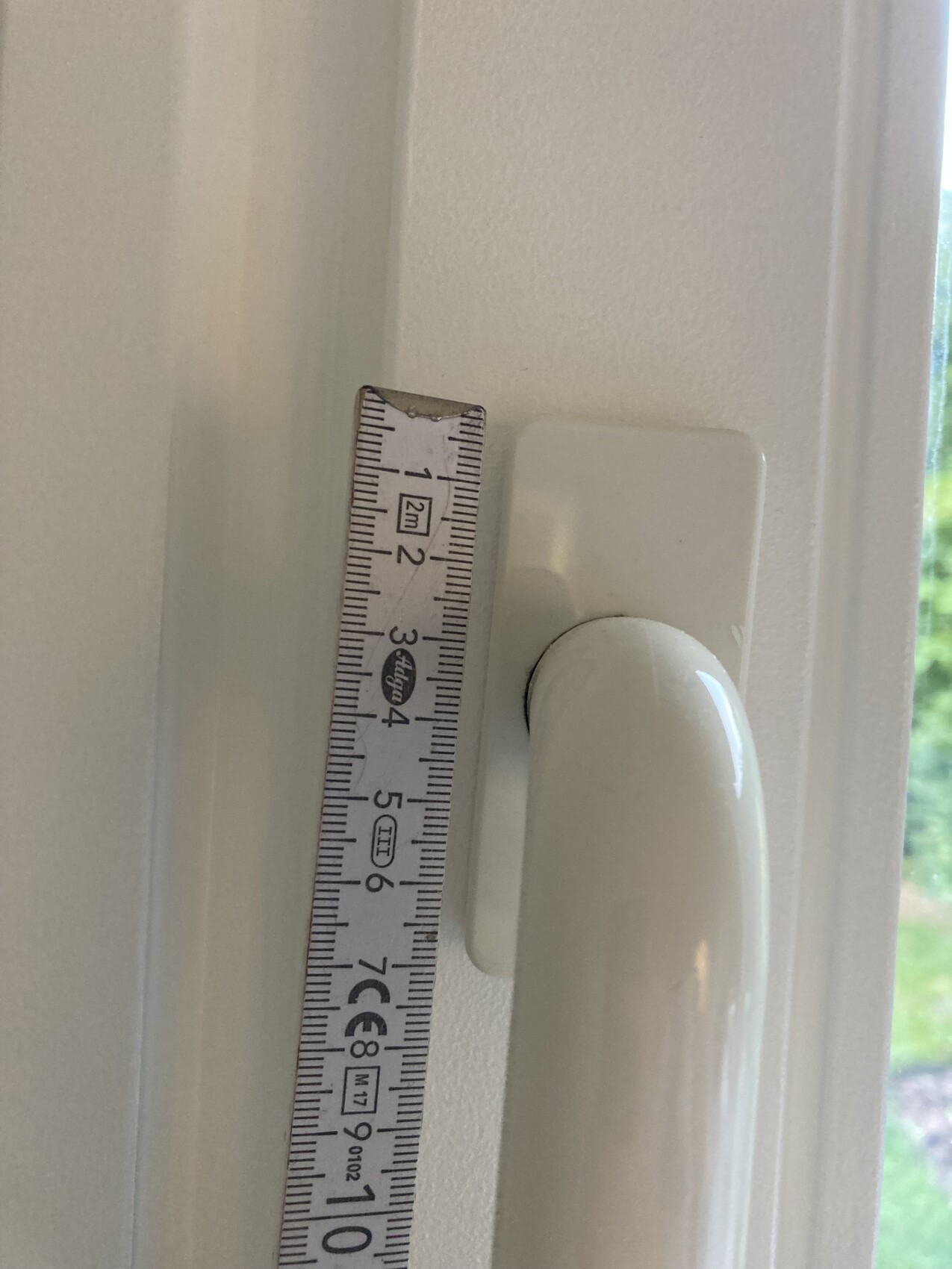 Close-up of a white window frame with handle, measuring tape beside to read the width.