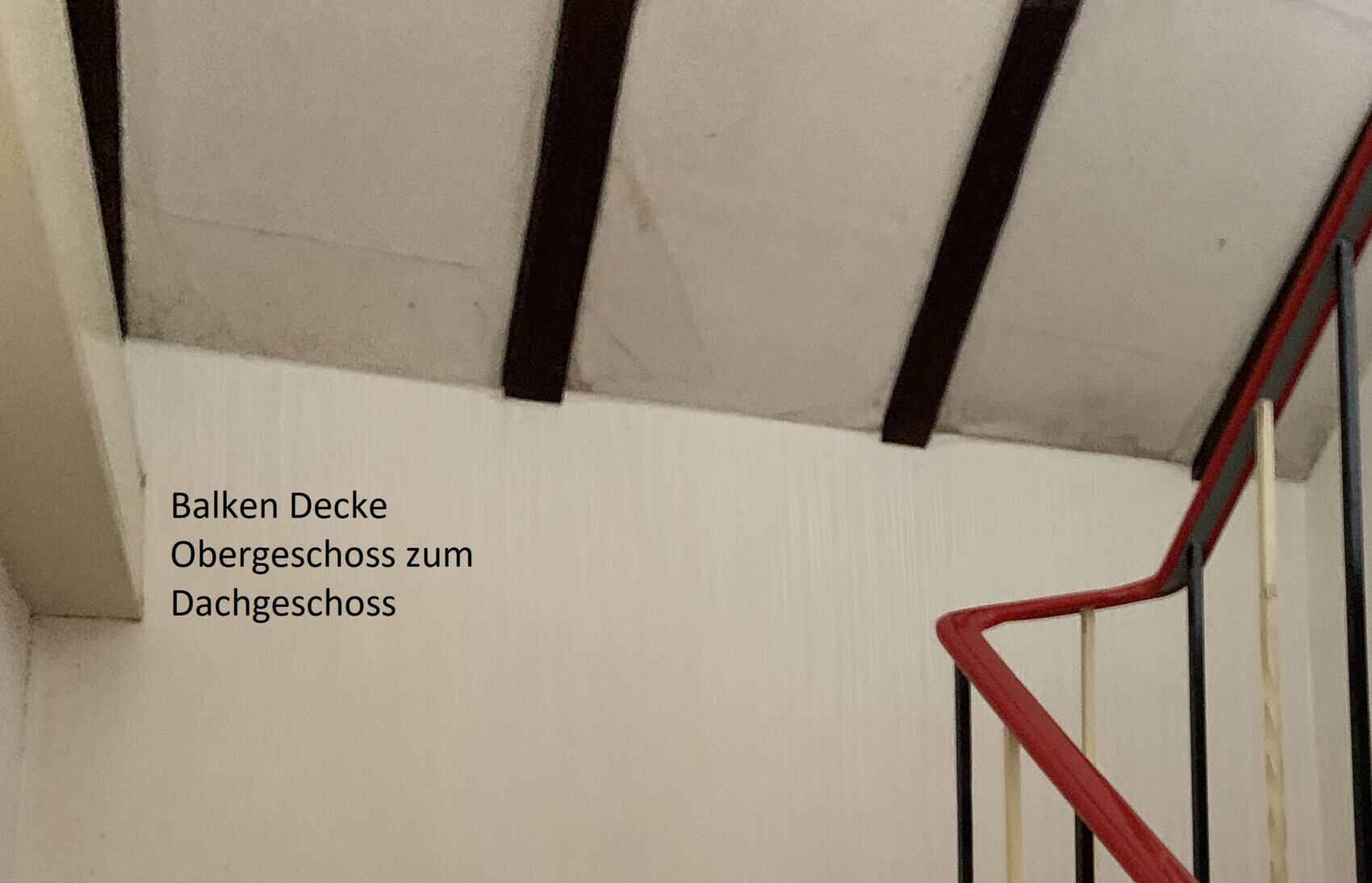Innenansicht eines Treppenhauses mit weißen Wänden, dunklen Deckenbalken und rotem Handlauf.