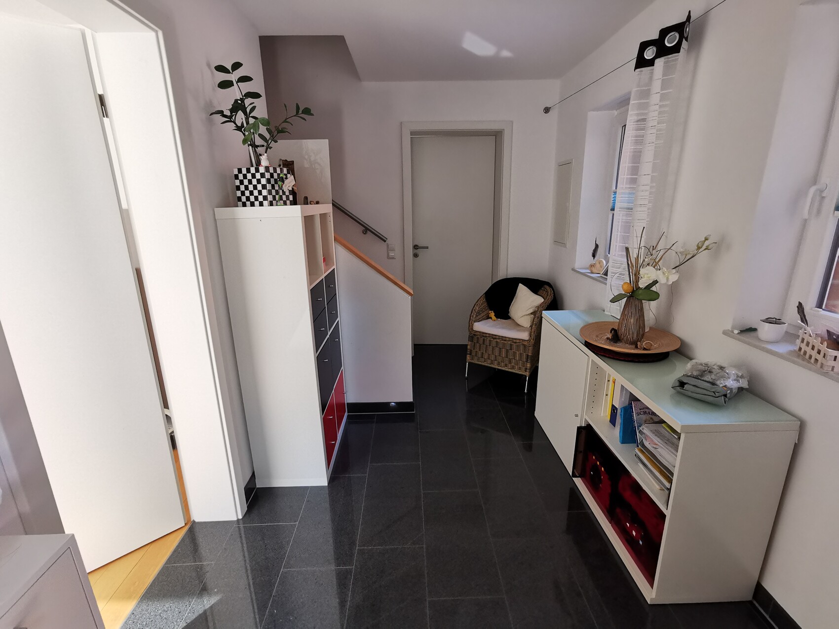 Bright hallway/entrance area with white furniture, plant, stairs, wicker chair, and window with curtain.