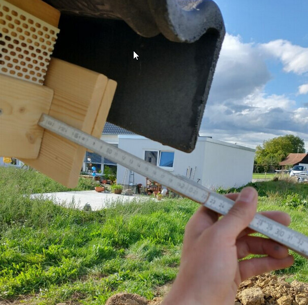 Person measuring the length of a roof tile on the house with a tape measure; garden in the background.