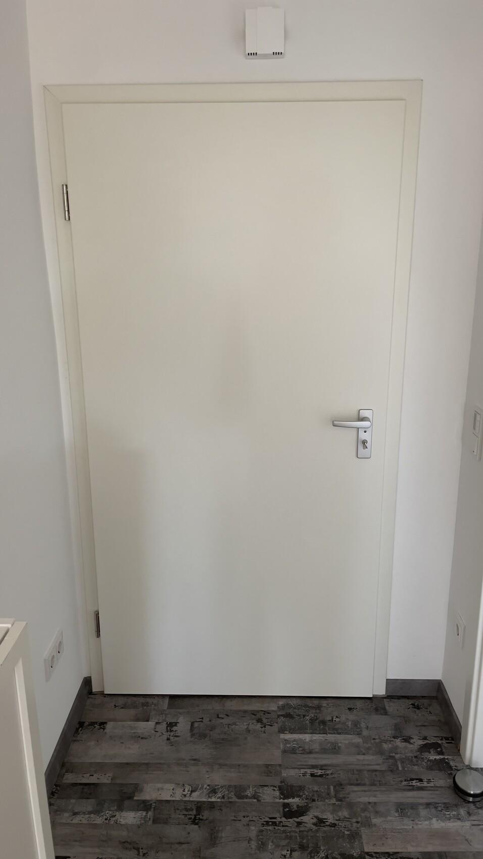 View of a white interior door in a hallway, handle on the right, dark floor tiles, wall fixture above the door.