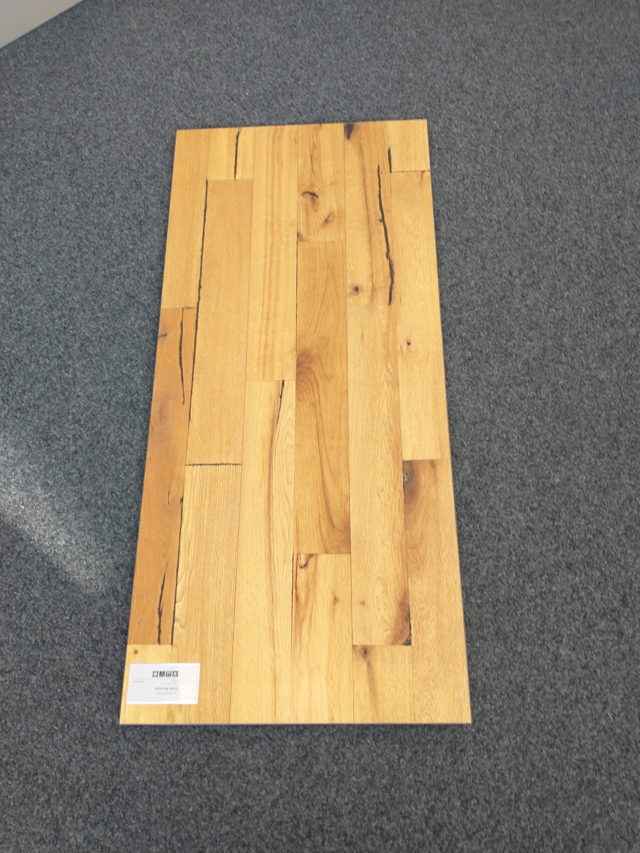 Wood plank floor sample in light boards on grey carpet, label in bottom left.