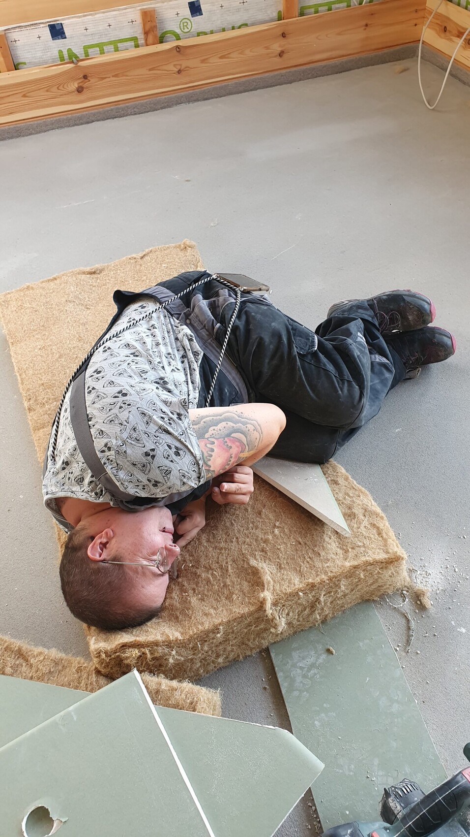 Man lying sideways on a mat at a construction site, with a rope around his upper body, tattoo on his arm.