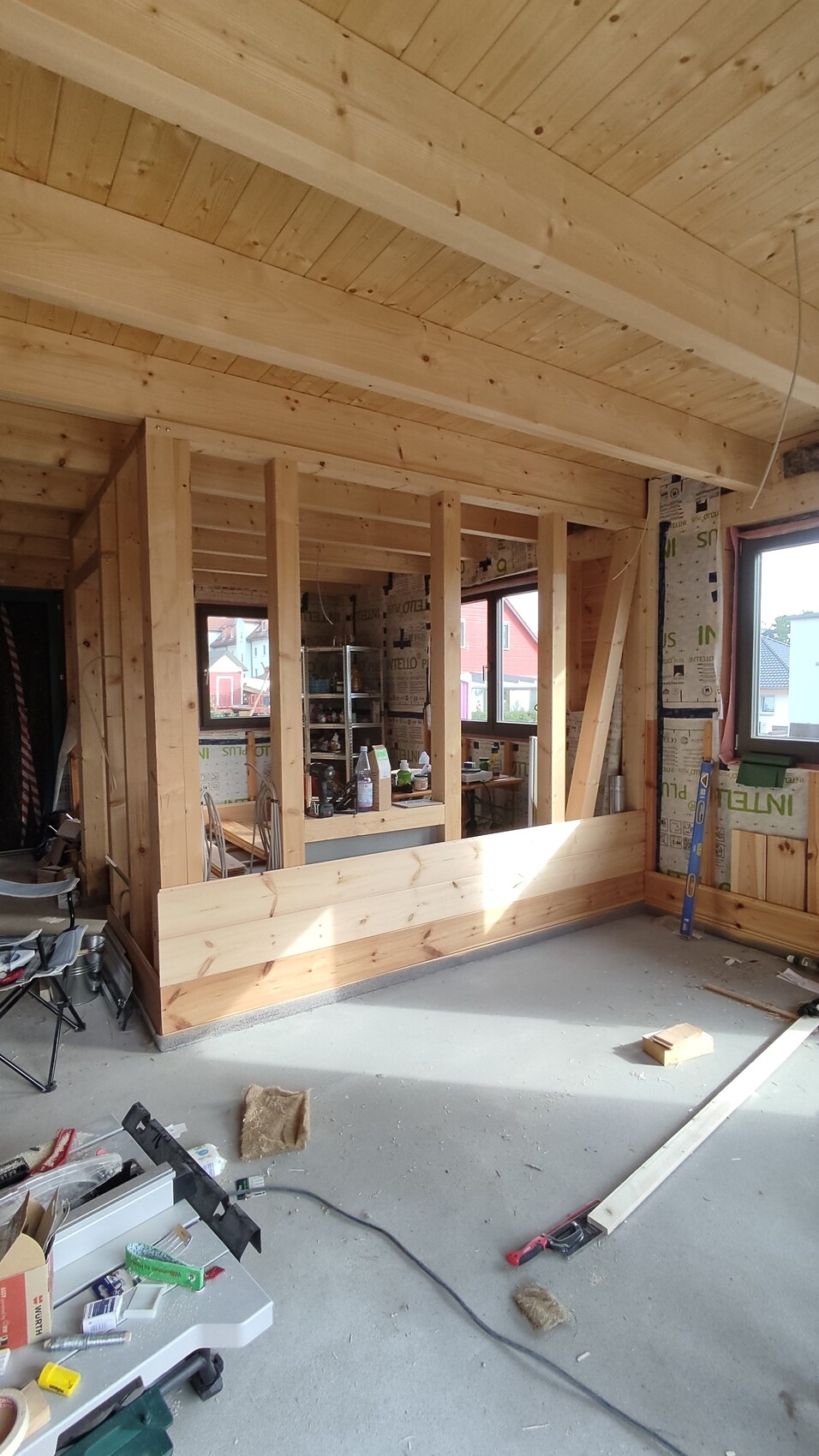 Shell construction of a wooden house: visible timber frame walls, ceiling beams, windows, tools.