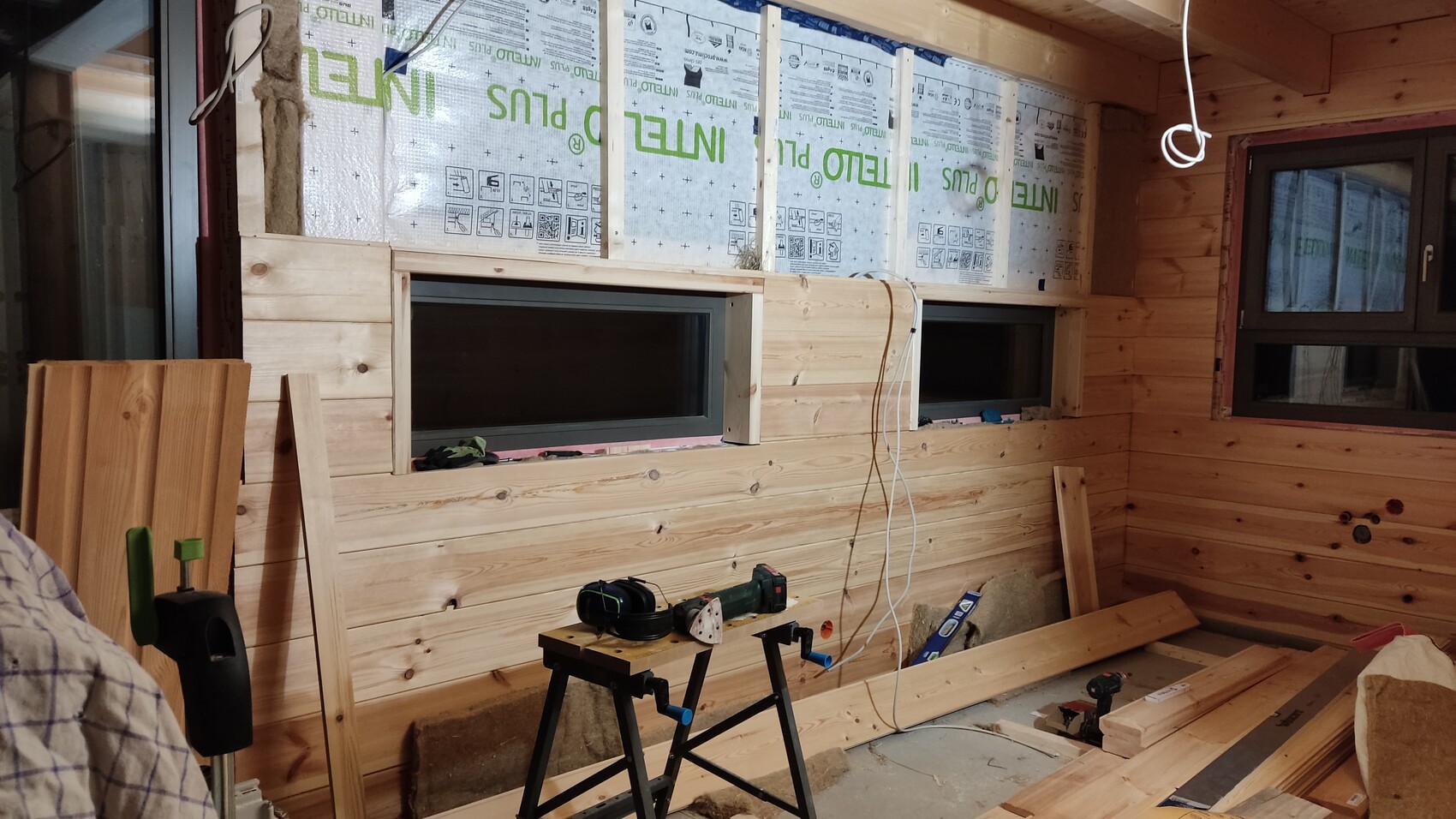 Interior of a wooden cabin under construction: wood cladding, two windows, tools and building materials.