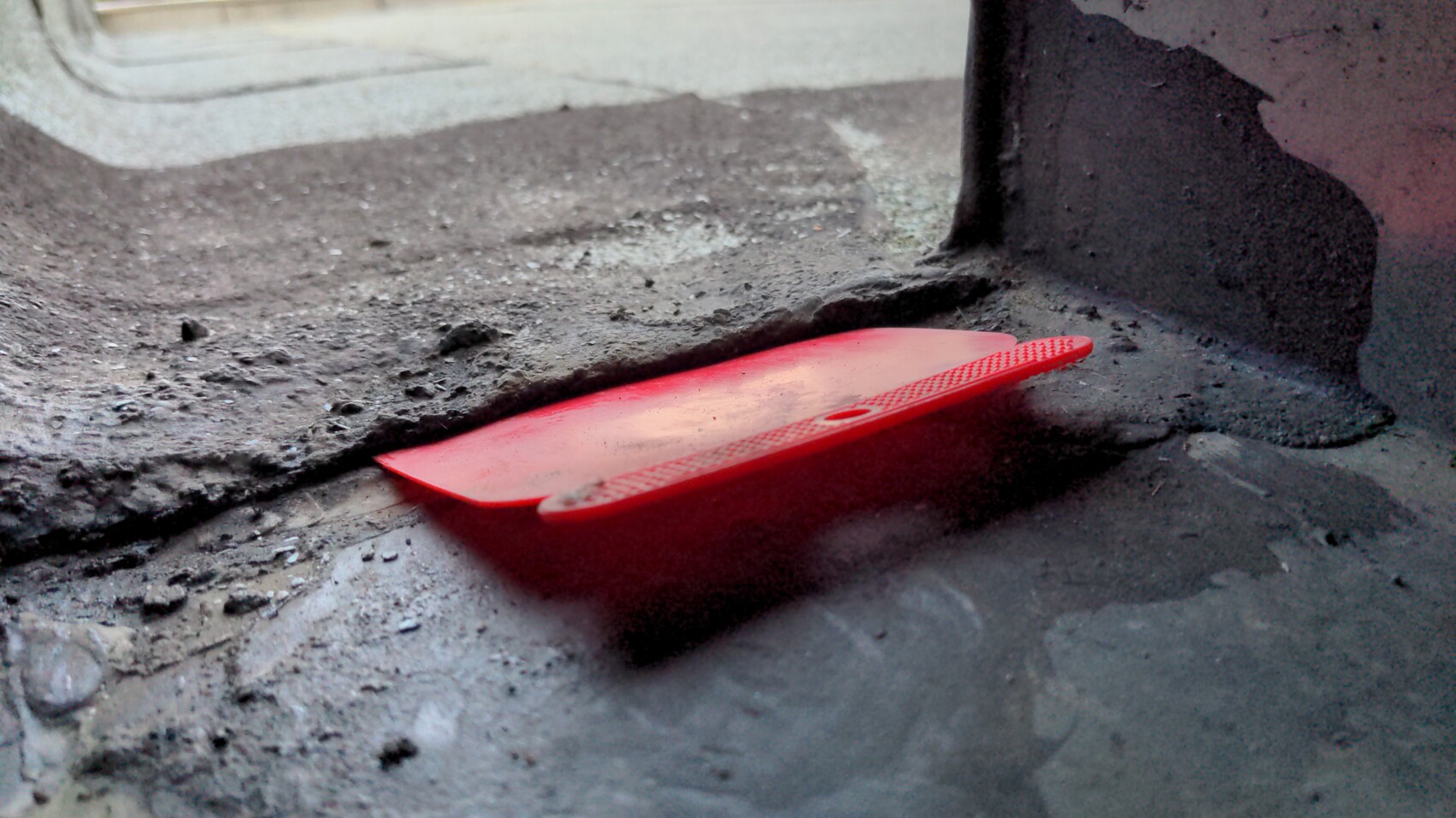 Red plastic scraper lying on dusty concrete floor next to a dark support.