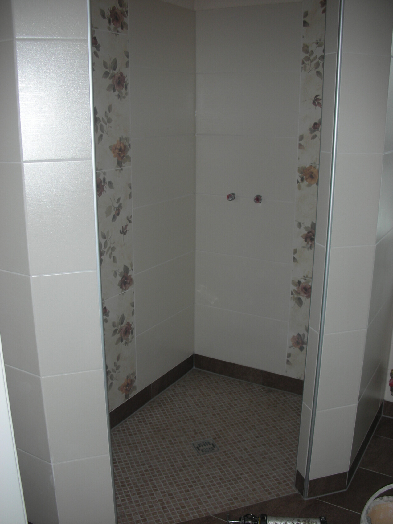 Corner of a shower cabin with floral tiles on the sides, brown floor mosaic, and faucets.