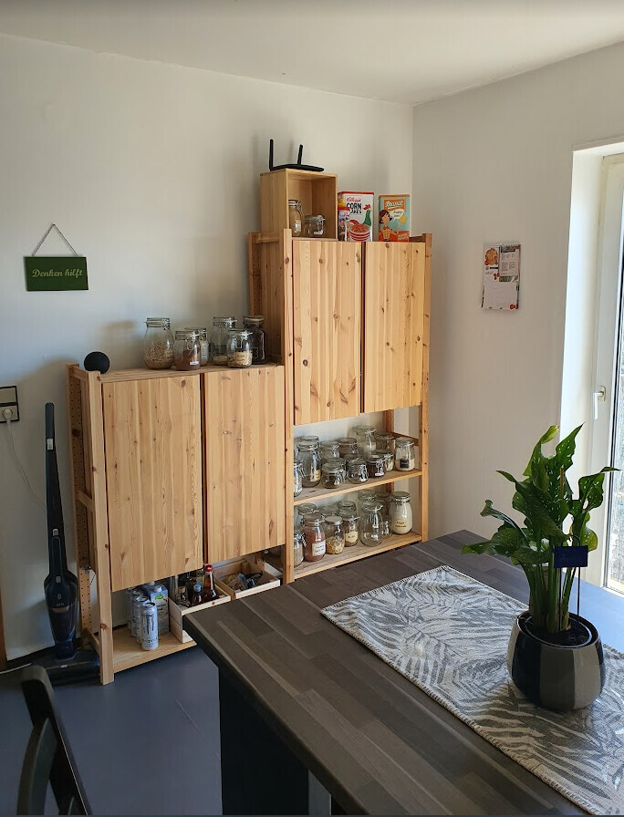 Wooden shelves with jars, storage containers and cereal boxes; dining table with a plant.