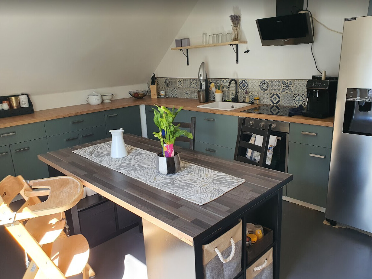 Modern kitchen with island, green base cabinets, wooden countertop, sink, coffee machine and plant.