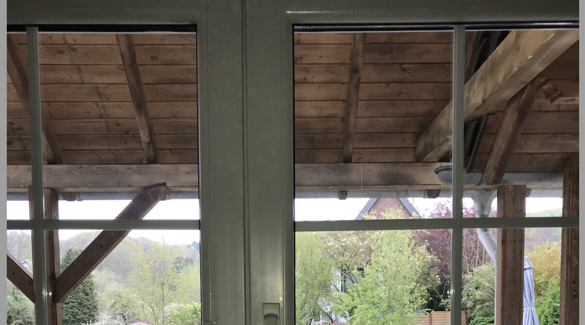 View through a glass door onto a wooden structure with roof beams and a garden with trees behind.