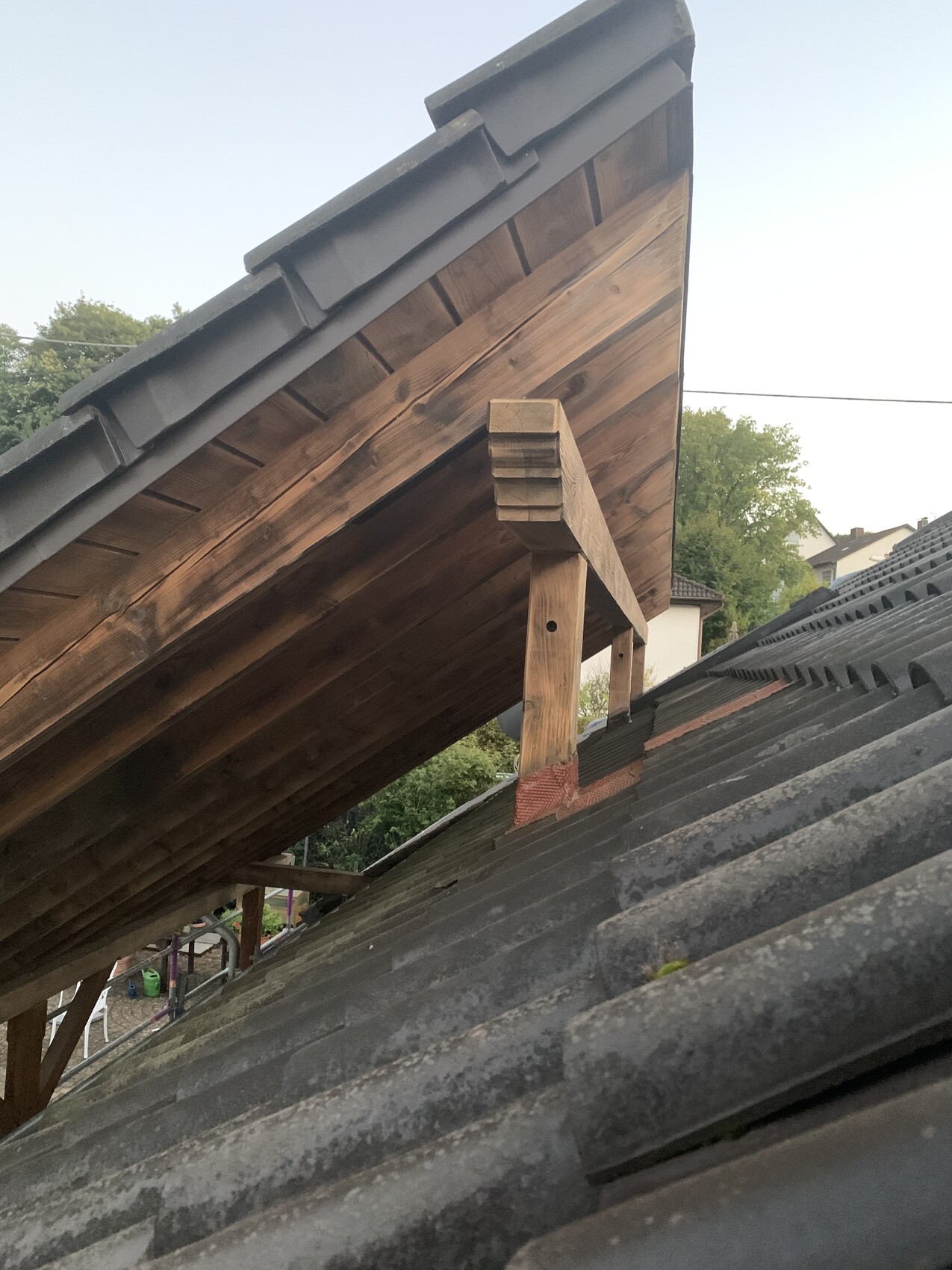 Sloped wooden roof with beam substructure and gray roof tiles; background with trees.