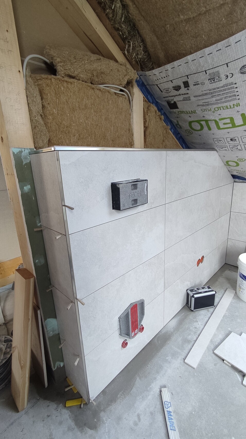 Construction site: wall with large light gray tiles, black electrical box, toolbox on the floor.