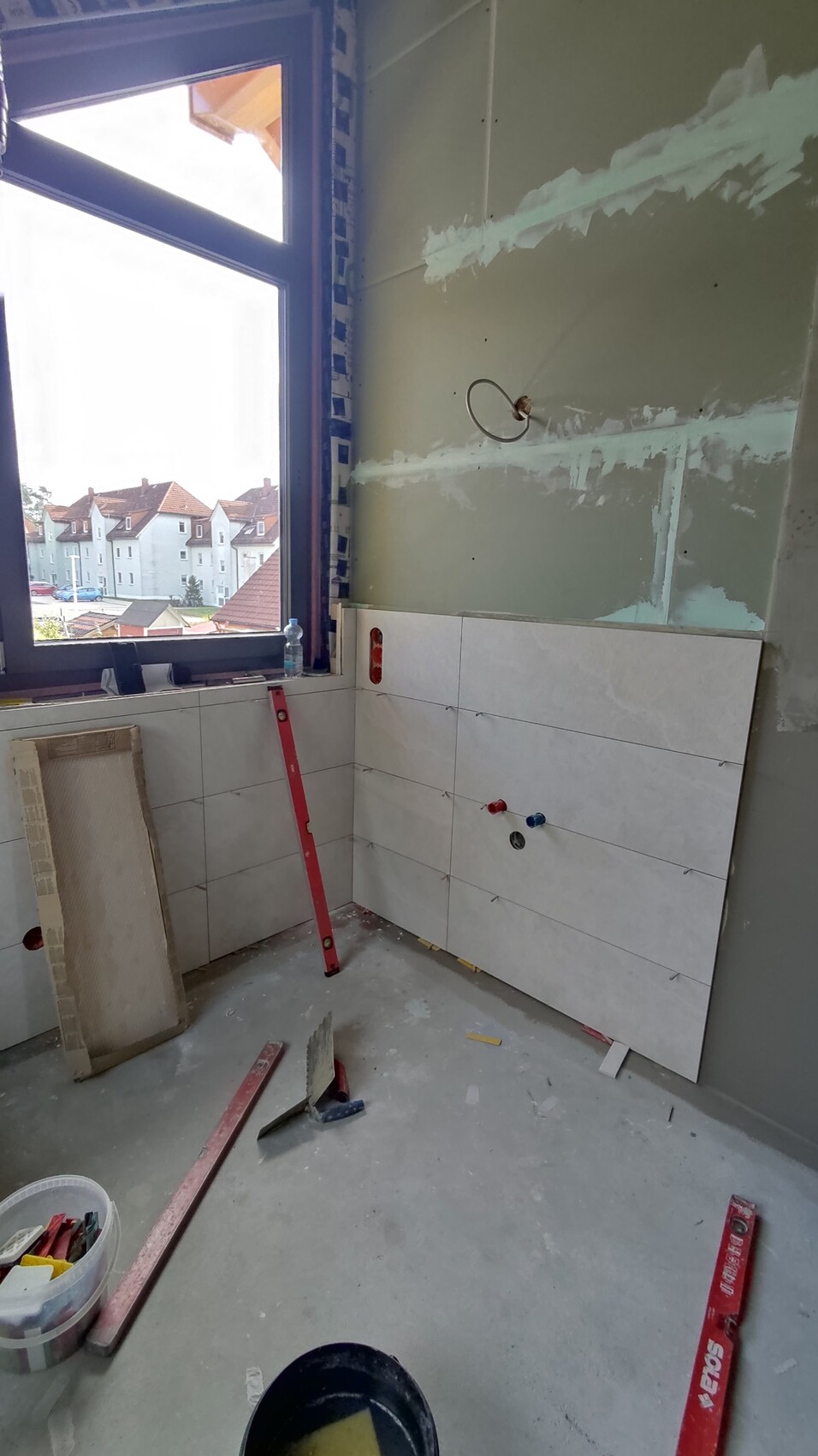 Renovation room: lower white tiles, window overlooking houses, building materials and tools.