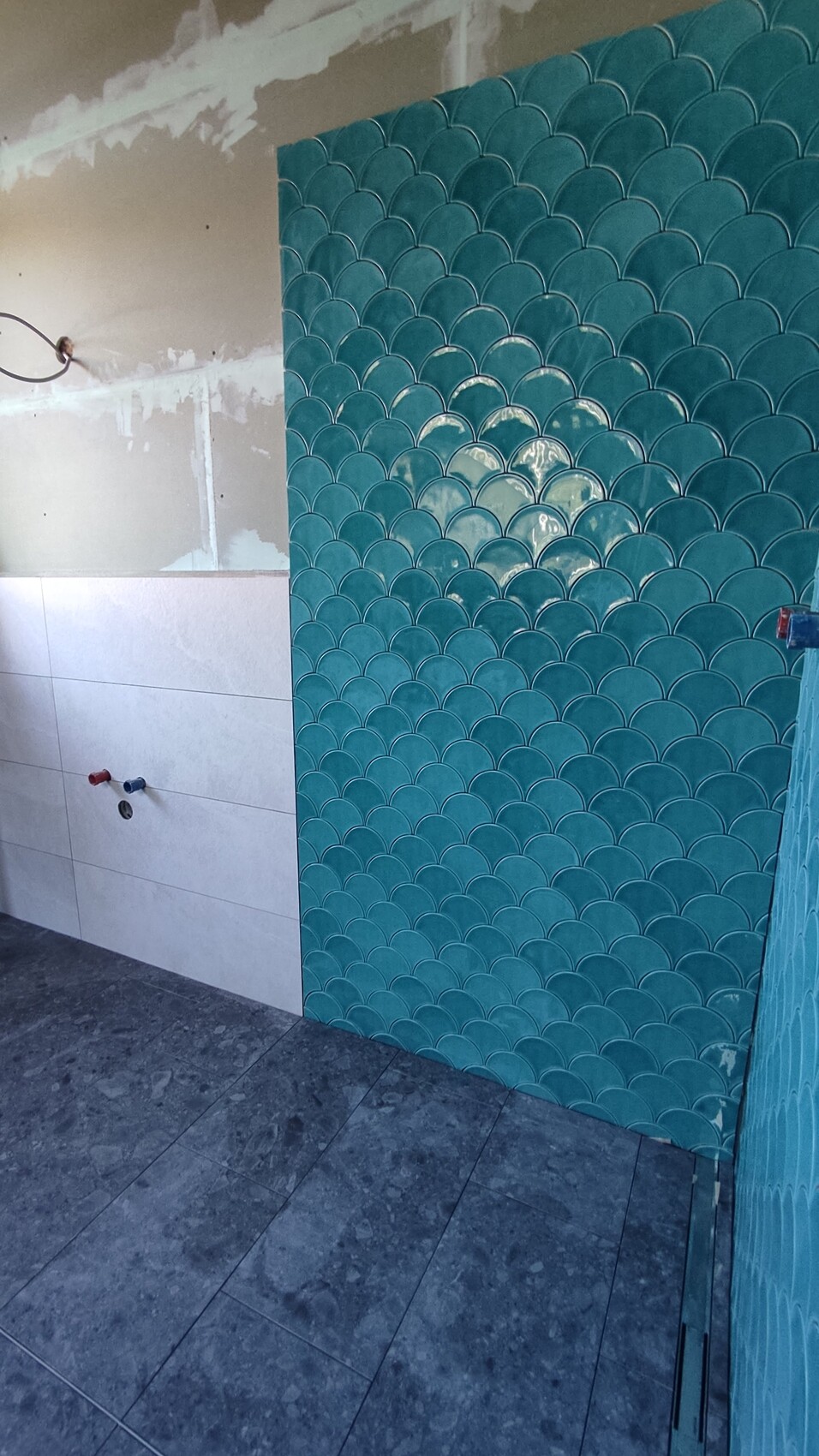 Bathroom wall with turquoise fish scale tiles; unfinished wall with pipe connections on the left.