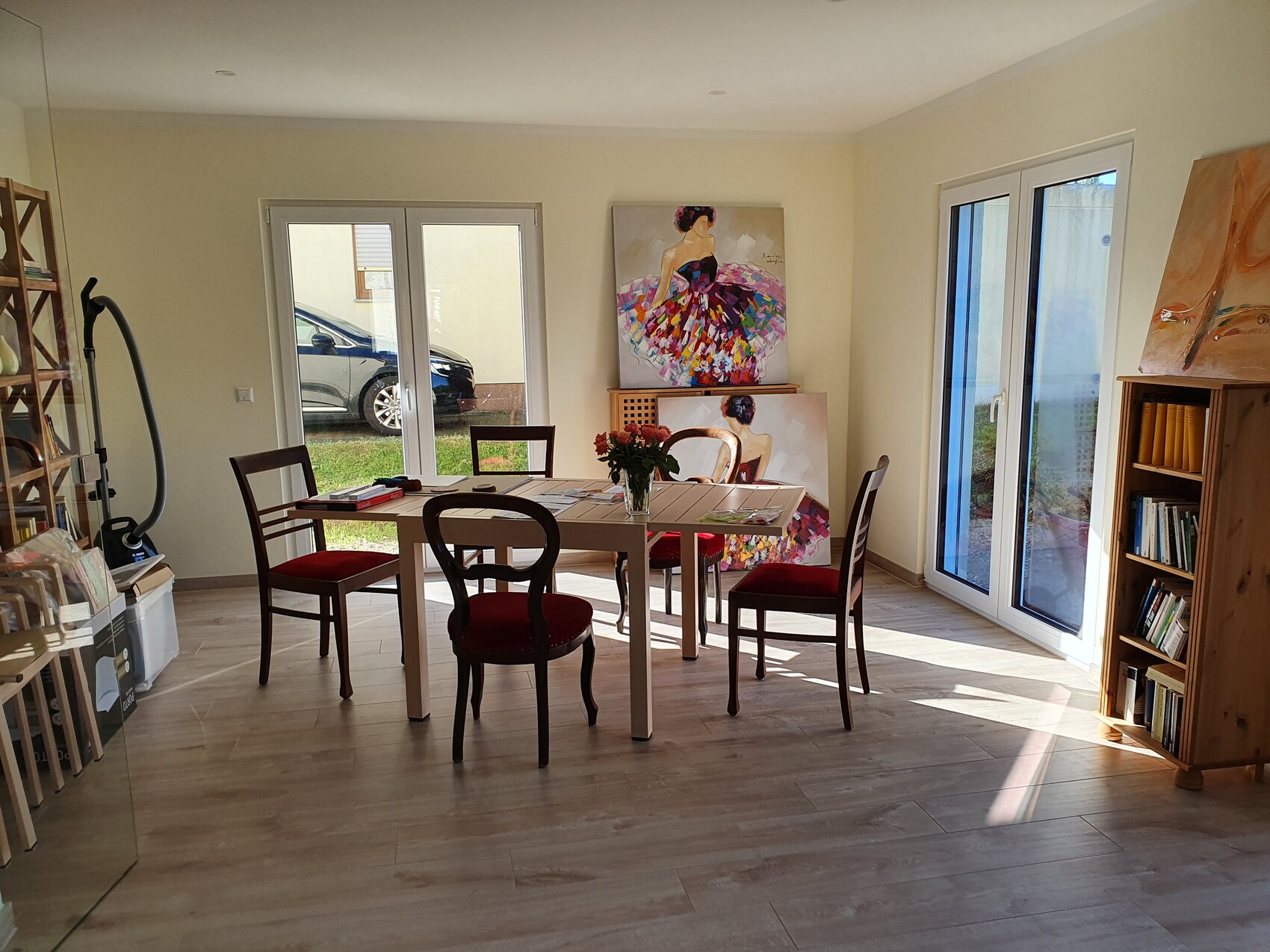 Bright dining and working room with dining table, four chairs, flower vase, paintings, and shelf.