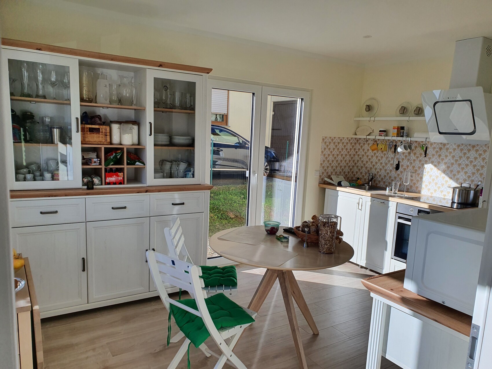 Bright kitchen with white cabinets, glass displays, round wooden table, and green seat cushions.