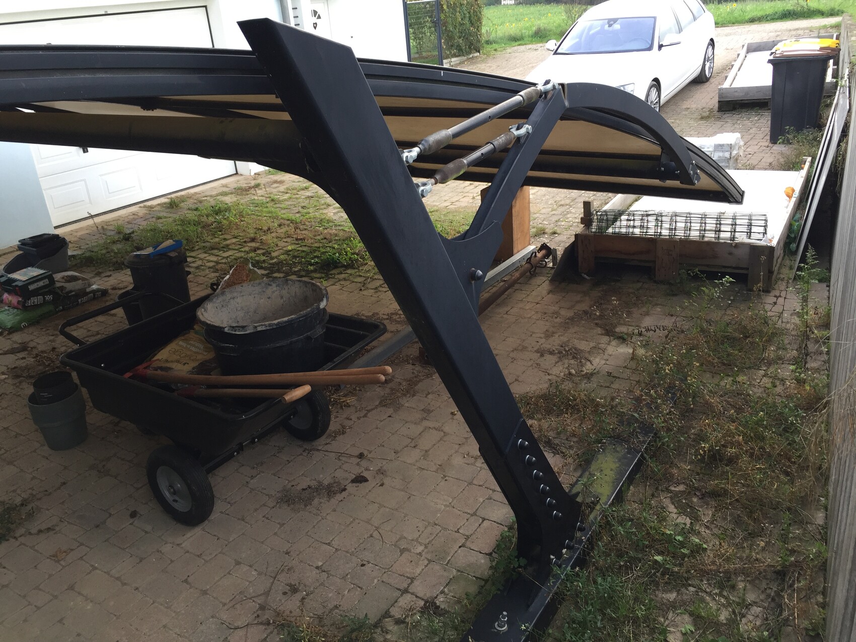 Large blue excavator arm in the yard, next to a wheelbarrow with buckets and tools; car in the background.