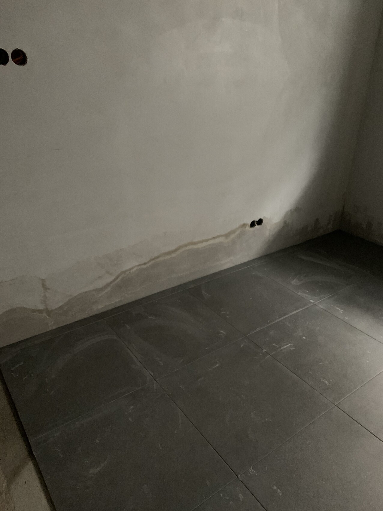 Corner of a damp room with cracked wall, water stains at the base and dark tiles.