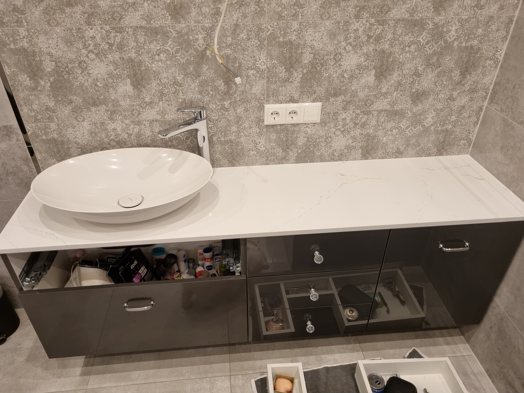 Modern washbasin countertop with round basin, chrome faucet, gray tiled wall, open cabinets.