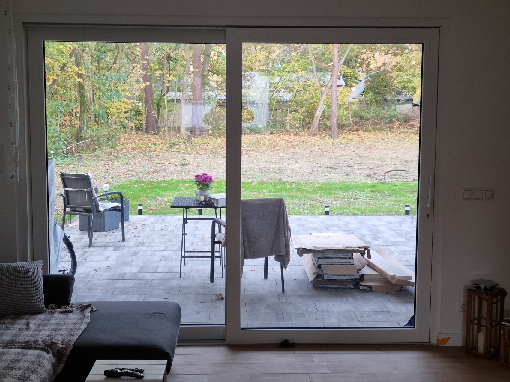 View through sliding door onto terrace with table, chairs, flowers, and stacked firewood in the garden.