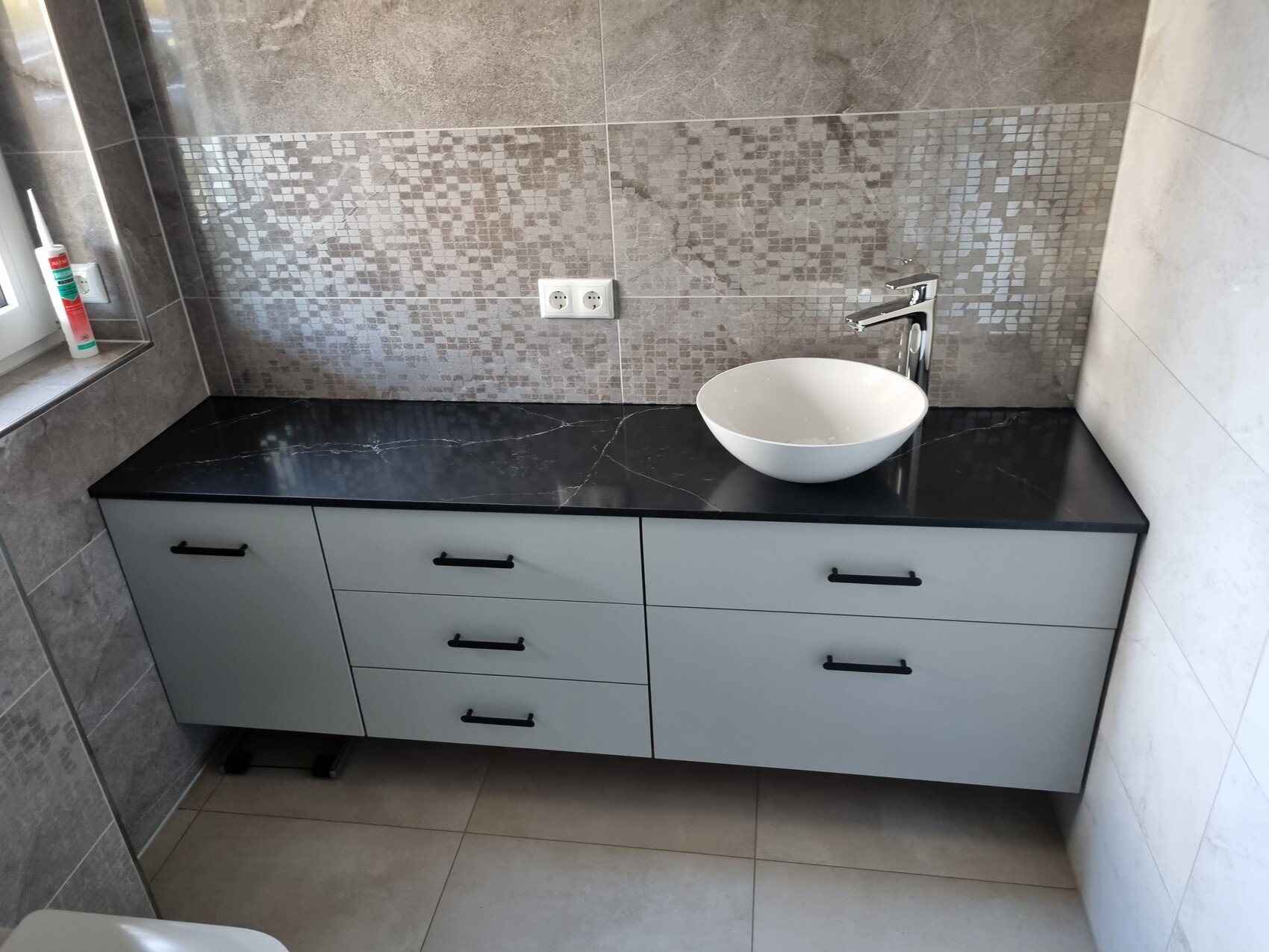 Modern bathroom washbasin with black countertop, round white sink, mosaic backsplash, chrome faucet.