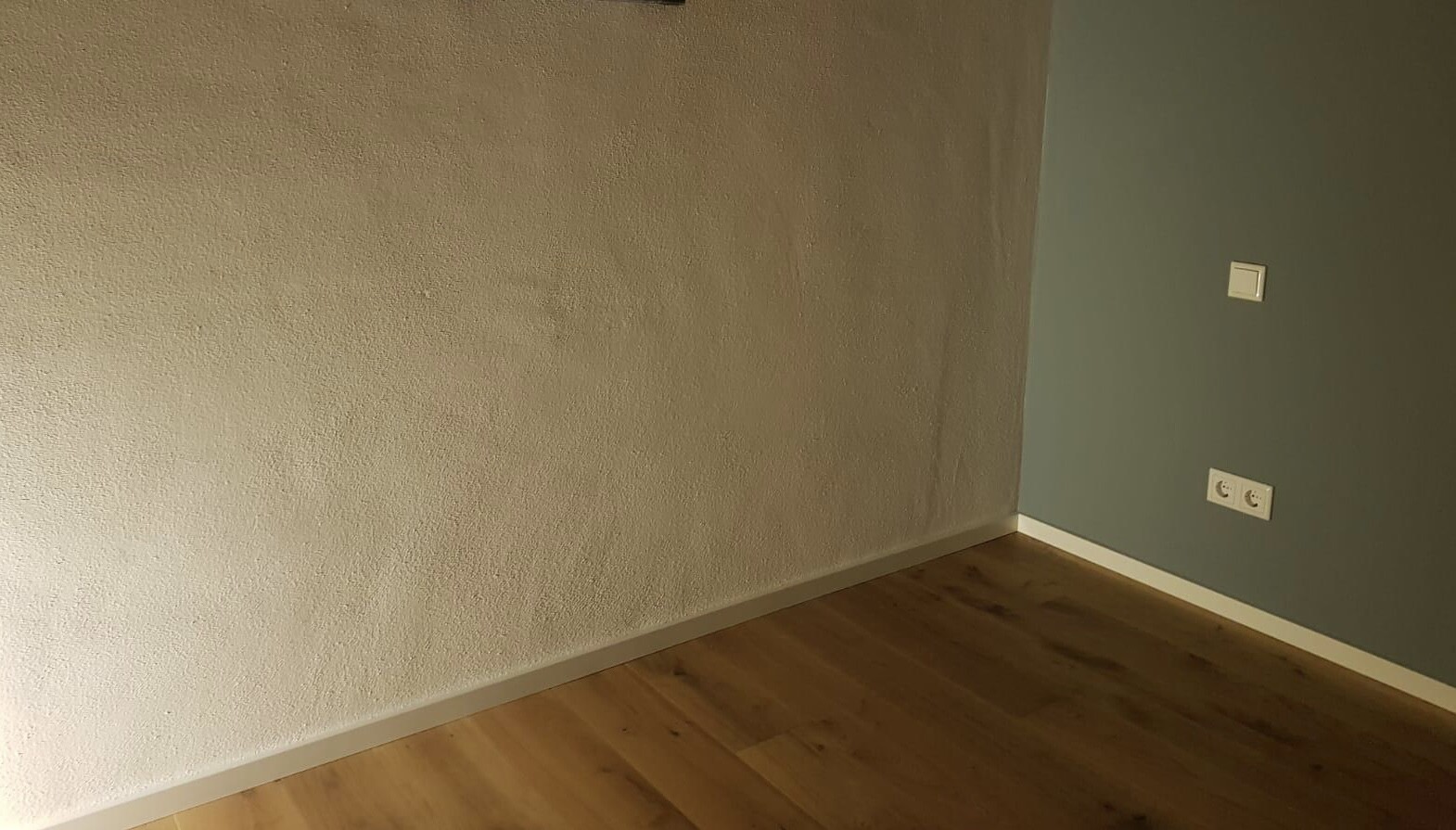 Corner room: left beige textured wall, right blue-green wall, wood floor, power outlets on the right wall.