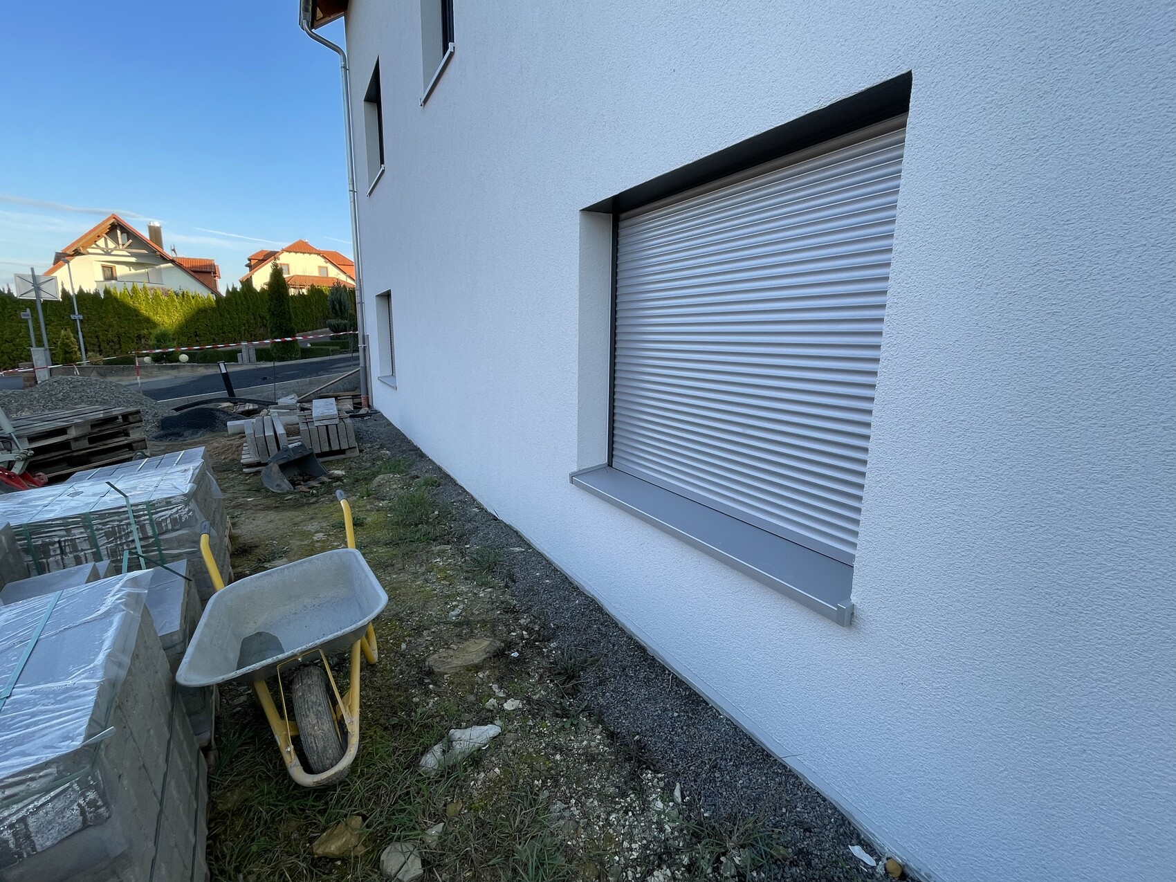 Exterior view of a white building with a closed roller shutter; building materials and wheelbarrows in front.