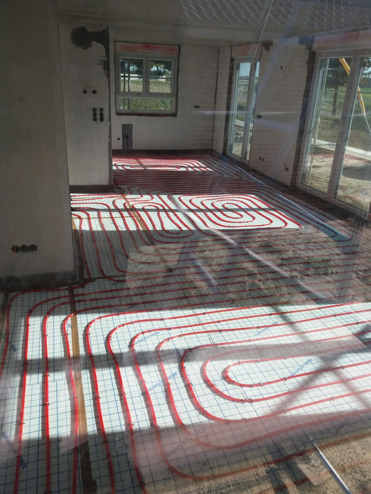 Underfloor heating: red pipes spiraled on mesh layer, indoor construction site with window walls.