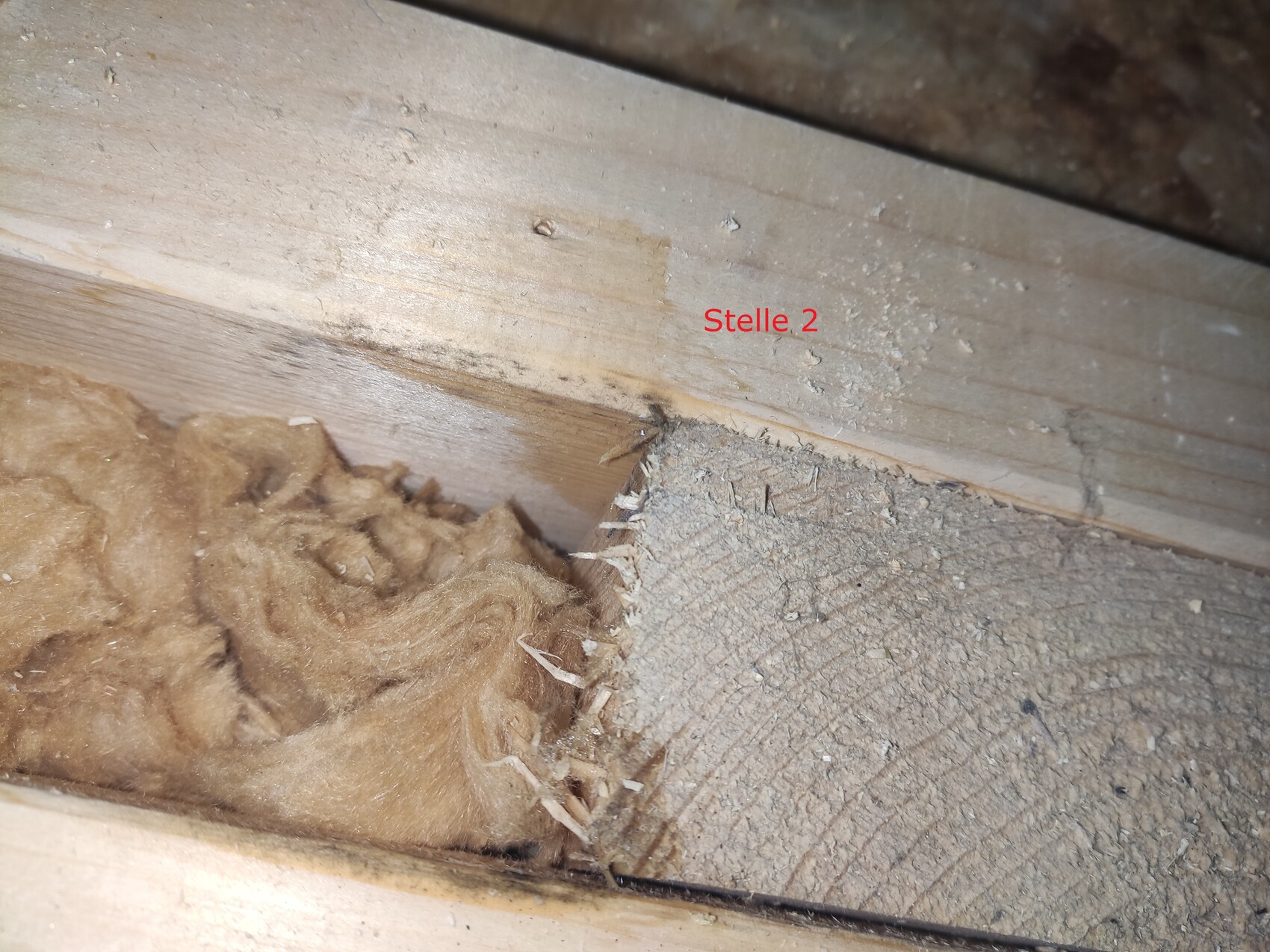 Dachboden: Spalt zwischen Holzbalken; braune Dämmwolle links, staubige Spanplatte rechts.