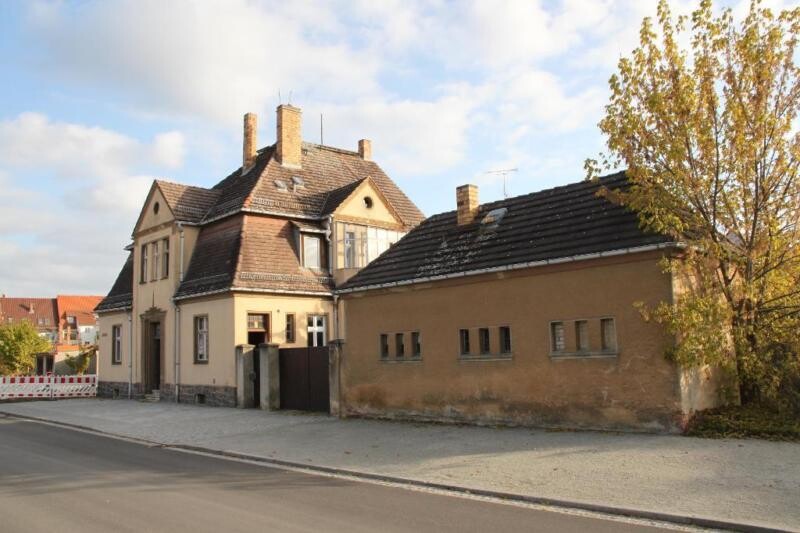 Zwei alte Häuser an einer Straße, beige Fassaden, Satteldächer, Baum mit Herbstlaub rechts