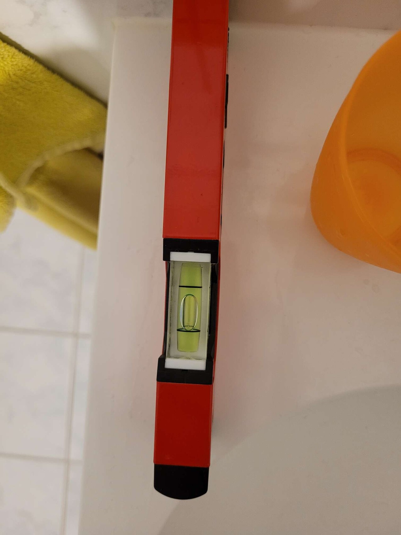 Red spirit level on white bathroom surface next to yellow towel and orange cup.