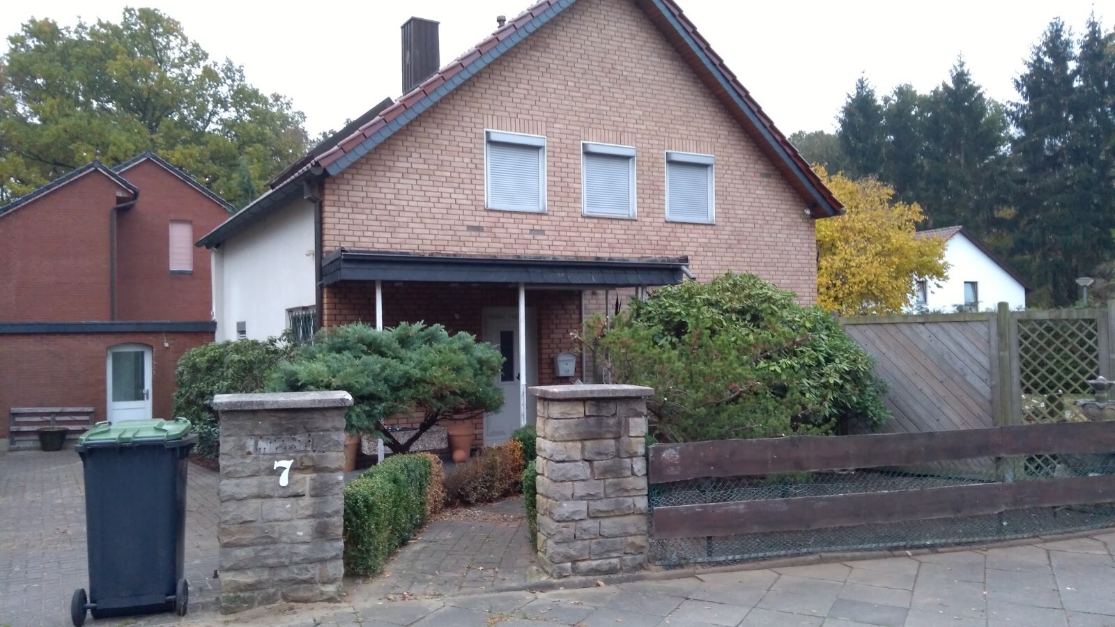 Front garden of a two-story brick house with three windows, stone pillar No. 7, trash bin.