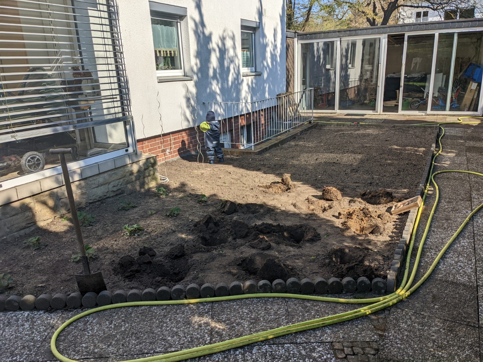 Child in blue work clothes watching earthworks in the backyard; spade, hose, and garden area.