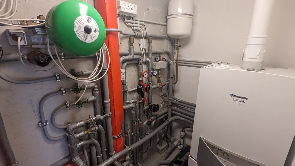 Heizungsraum mit vielen Rohren, grünem Ausdehnungstank und Boiler rechts.