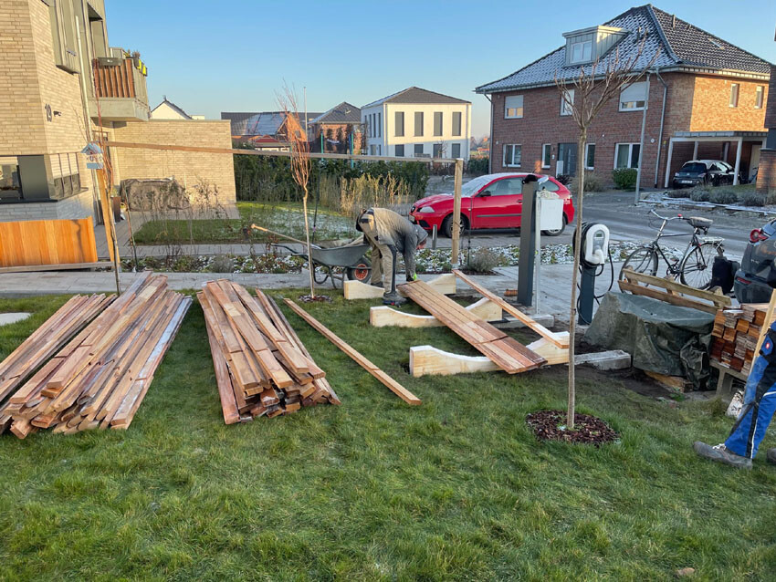 Person working on wood in the front yard; stack of wooden boards, construction timber, residential houses in the background.