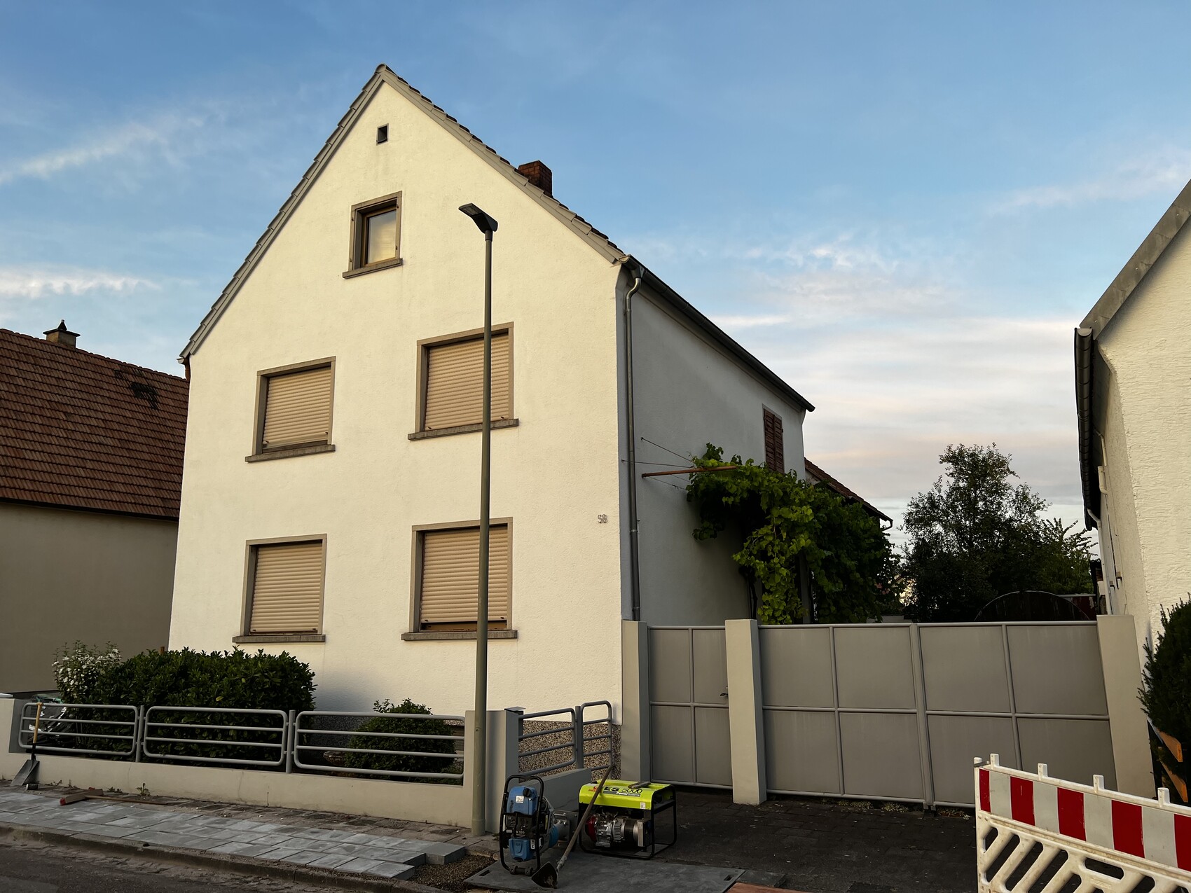 Weißes mehrstöckiges Haus mit Giebeldach, Rollläden, Straßenlampe, Bauzaun und Generator.