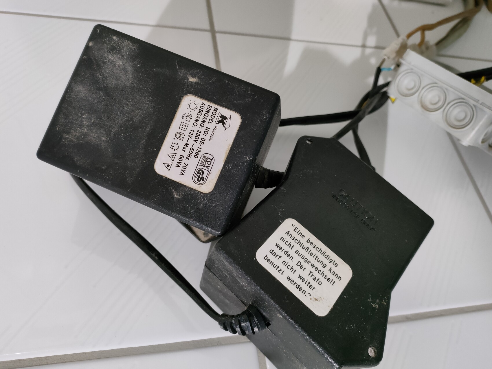 Zwei dunkle Netzteile/Transformatoren liegen auf weißem Fliesenboden, Kabel verbunden.