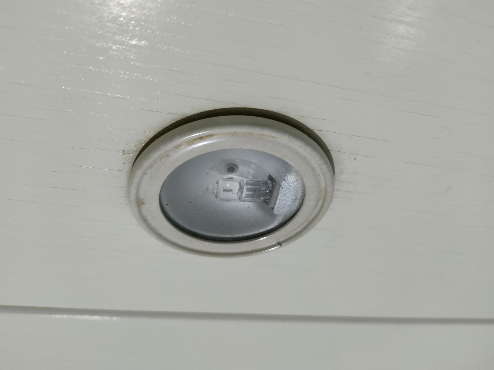 Runder Deckenstrahler mit sichtbarer Glühbirne in silbernem Metallrahmen an weißer Decke.