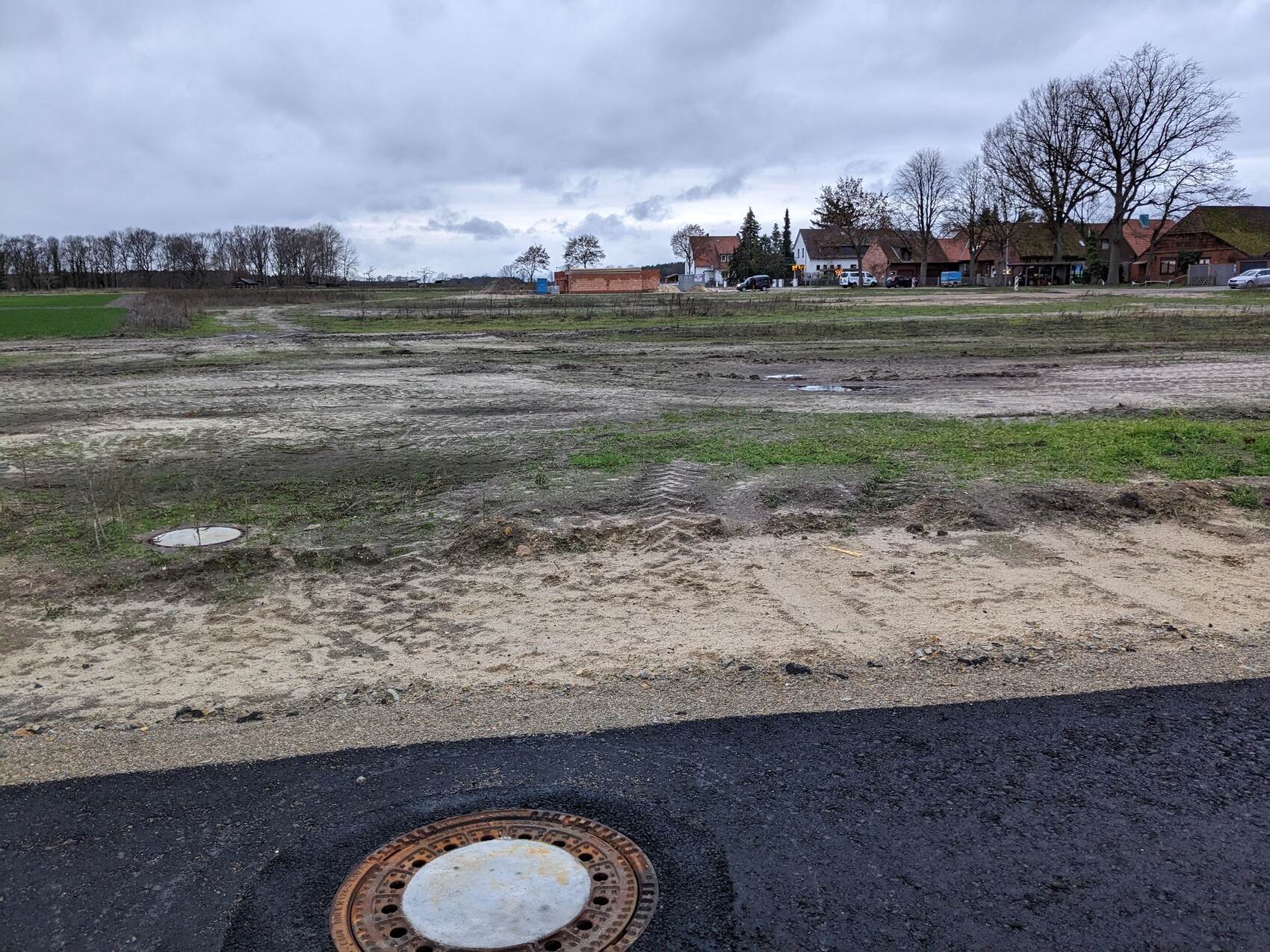 Open construction site with mud, grass, houses in the background and manhole cover in the foreground